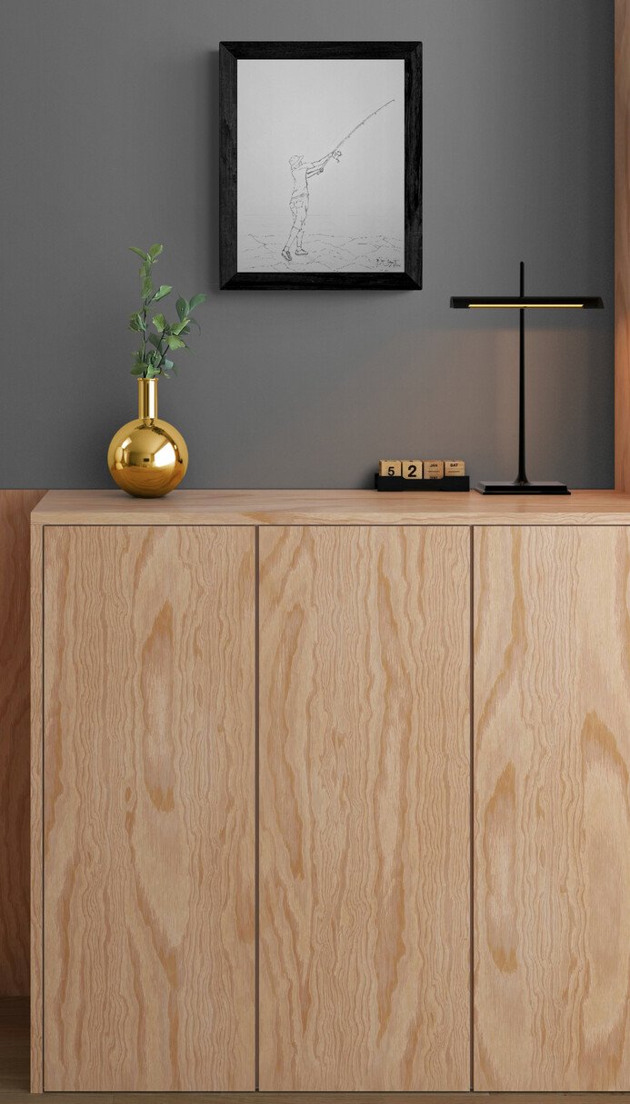 Fisherman No. 3 original drawing by Raul Miguel Sarante displayed in a black frame on a gray wall above a light wood cabinet, complemented by a gold vase and modern black table lamp.